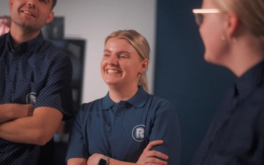 Professionelt stillbillede fra en videoproduktion til en brandingfilm, der fanger en smilende medarbejder i en autentisk samtale med kolleger. Dette eksempel på erhvervsvideo illustrerer brugen af visuel storytelling til at menneskeliggøre brands og skabe engagerende content af høj teknisk kvalitet i et moderne arbejdsmiljø.