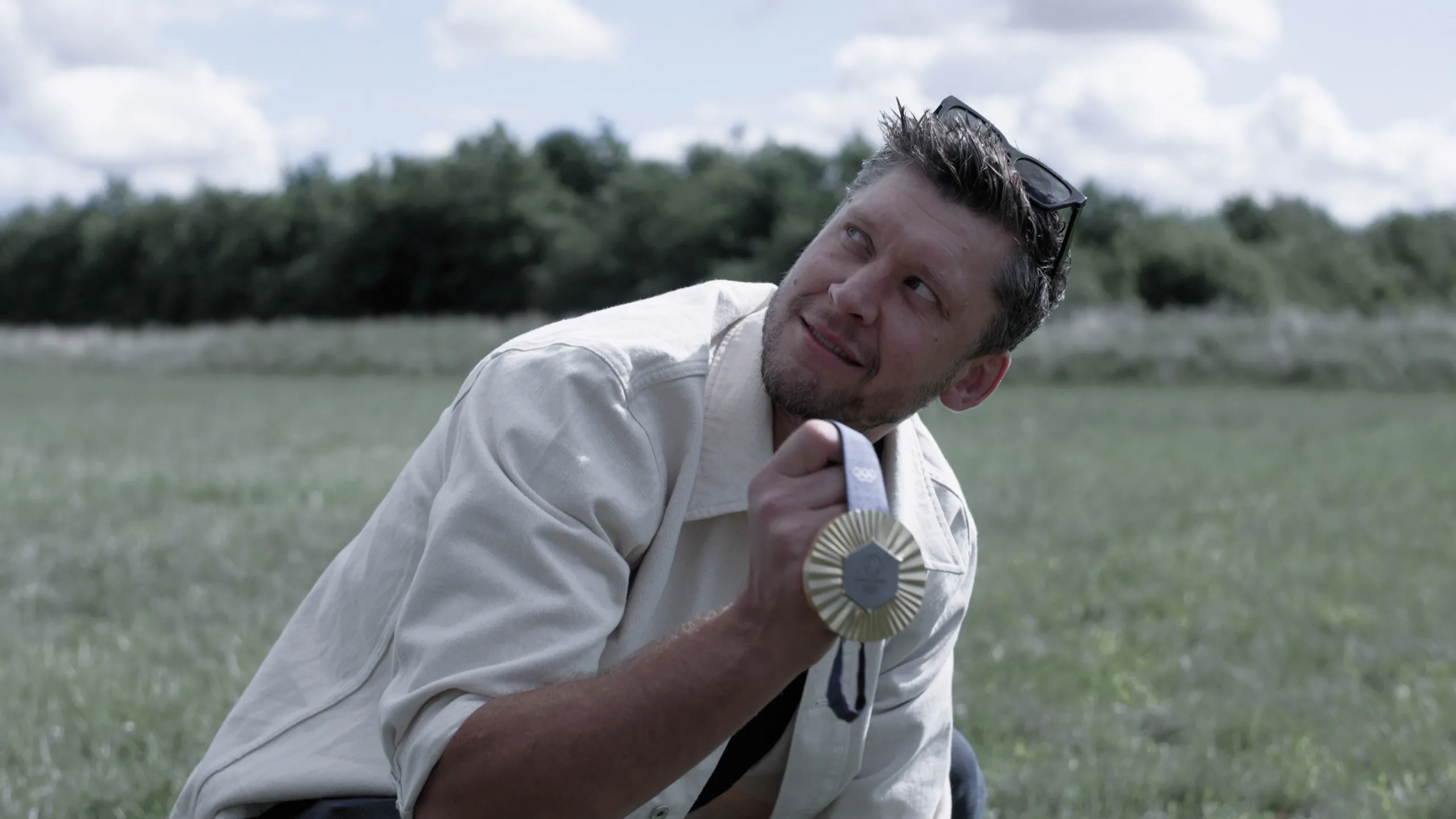 Cinematisk stillbillede fra en professionel videoproduktion, der viser en smilende mand, som holder en guldmedalje i et grønt udendørs landskab. Billedet er et eksempel på visuel storytelling og produktion af brandingfilm, der effektivt formidler følelser, succes og stærke personlige historier gennem video-content i høj kvalitet.