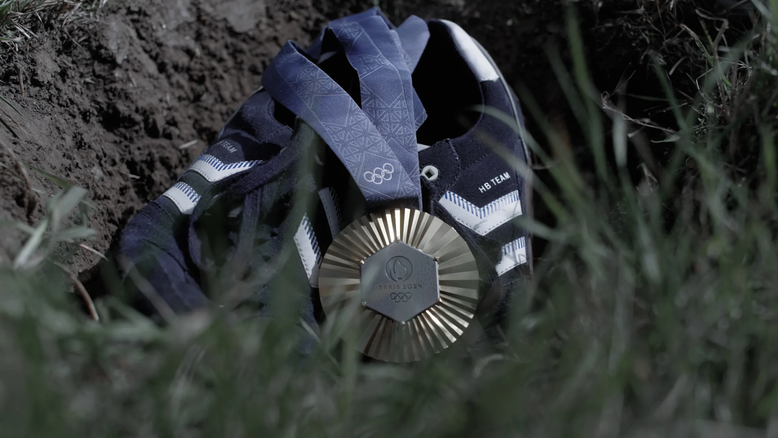 Cinematisk stillbillede fra en professionel videoproduktion, der viser en guldmedalje fra Paris 2024 og et par mørkeblå sportssko placeret i et hul i jorden. Billedet demonstrerer avanceret visuel storytelling og symbolik i en brandingfilm, med fokus på detaljerig komposition og professionel color grading.