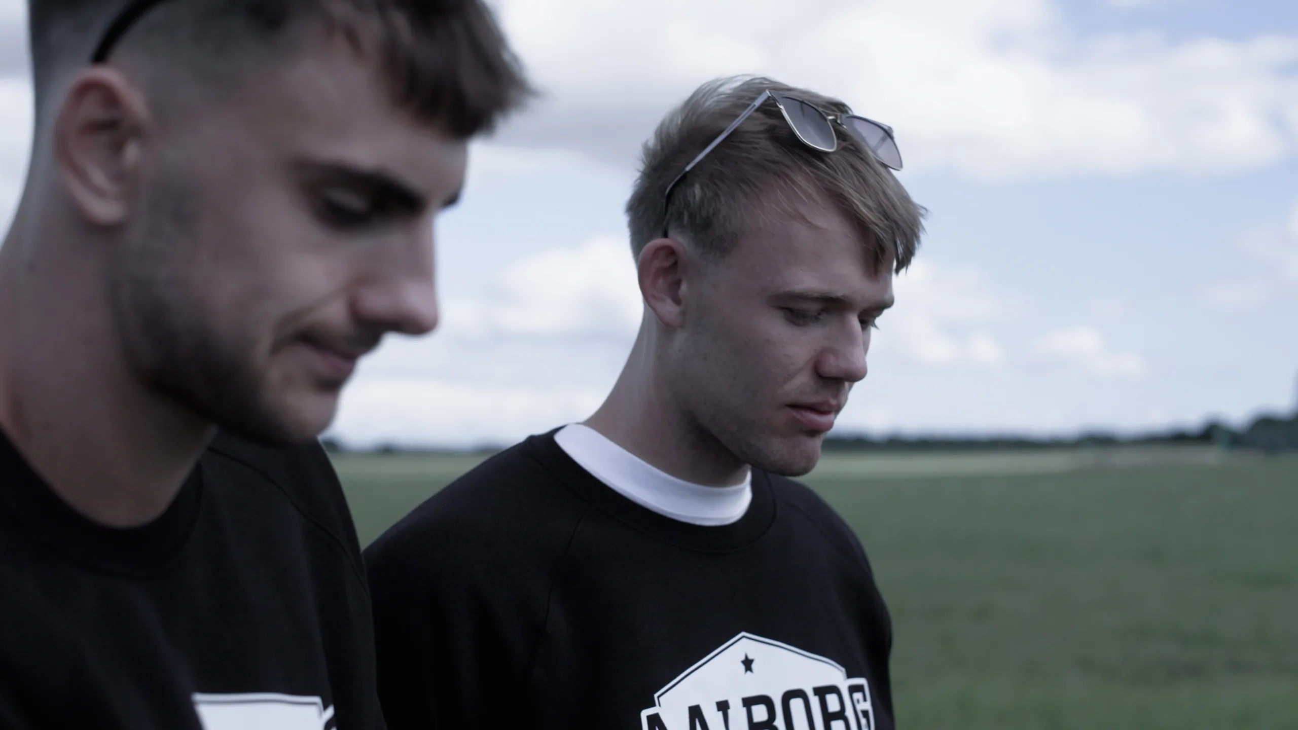 Cinematisk stillbillede fra en professionel videoproduktion, der viser to mænd i sorte sweatshirts med logo i et lyst udendørs miljø. Dette tæt-på klip demonstrerer ekspertise inden for visuel storytelling og produktion af brandingfilm, der skaber en autentisk og engagerende stemning for virksomheder.